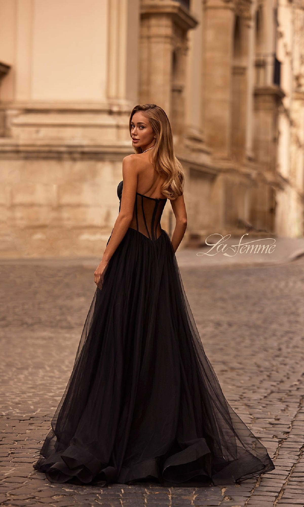 A woman in the La Femme 32771 black strapless long prom dress stands on a cobblestone street, glancing over her shoulder in an urban setting.